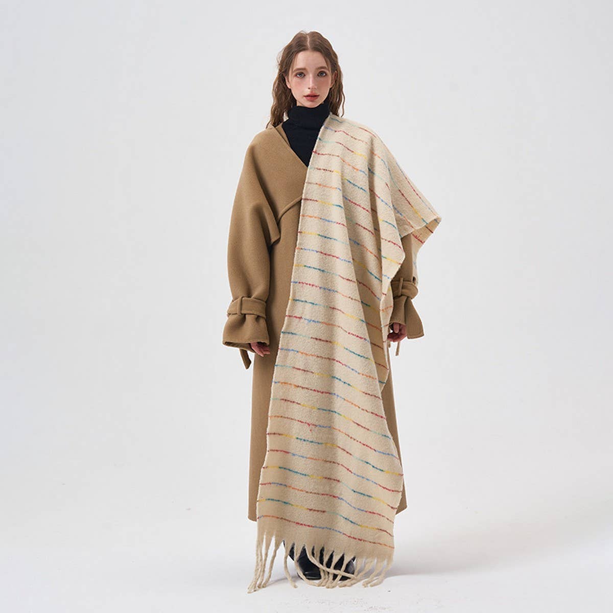 A model stands against a plain white studio background wearing a long tan coat over a black turtleneck, with a large cream shawl draped over her shoulders. The shawl has thin multicolored horizontal stripes and fringe at the bottom.