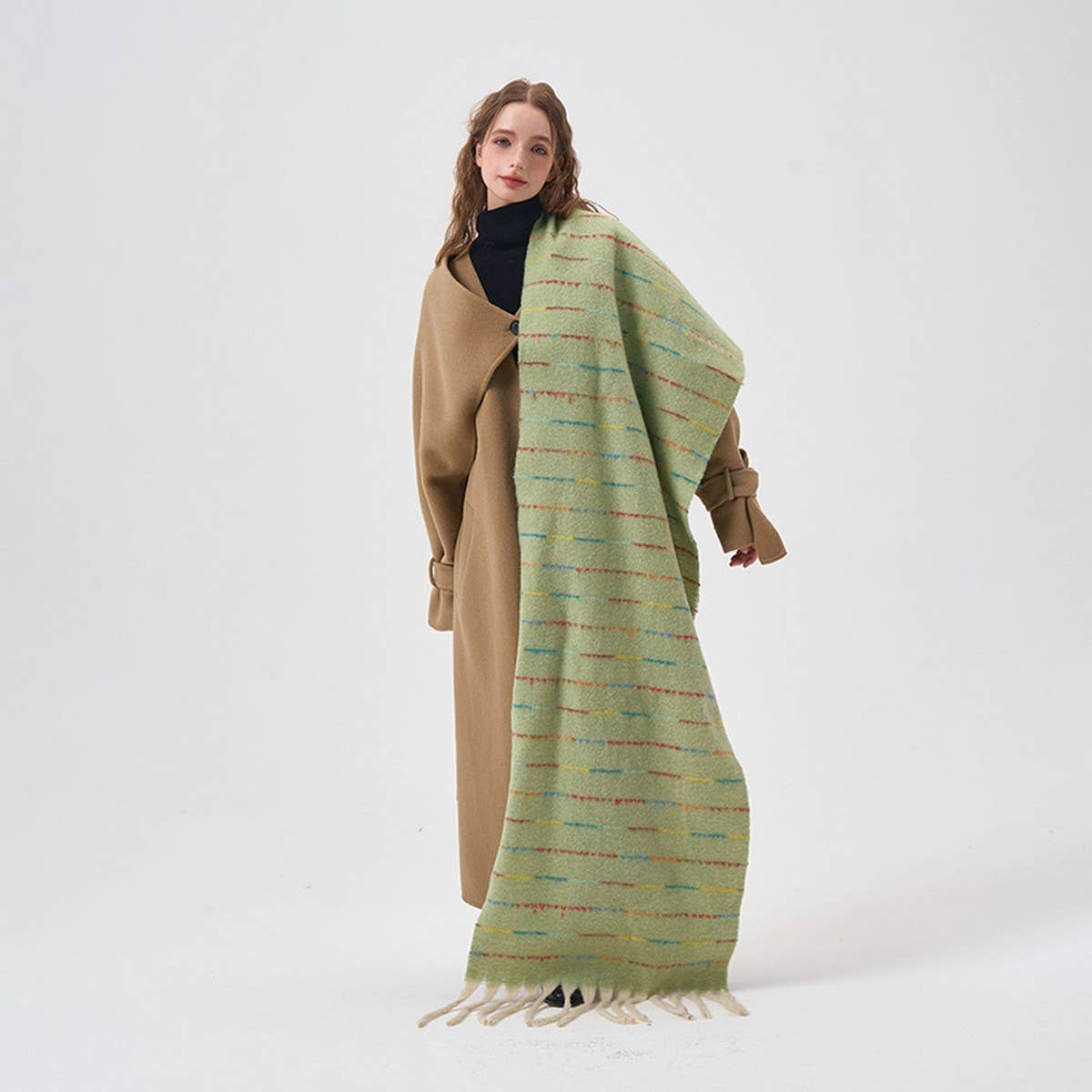 A woman stands against a plain light gray studio background wearing a long tan coat over a black turtleneck, with a large pale green patterned wrap draped over her shoulders and hanging down to the floor.