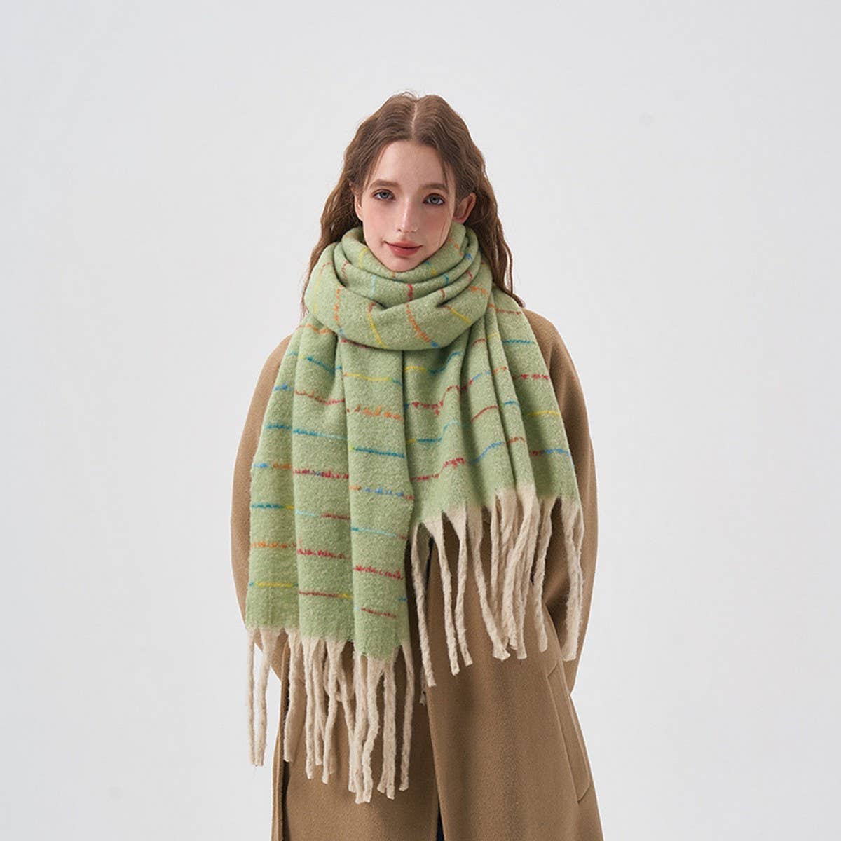 A woman wearing a light green plaid scarf with cream fringe, styled over a tan coat against a plain light gray background.