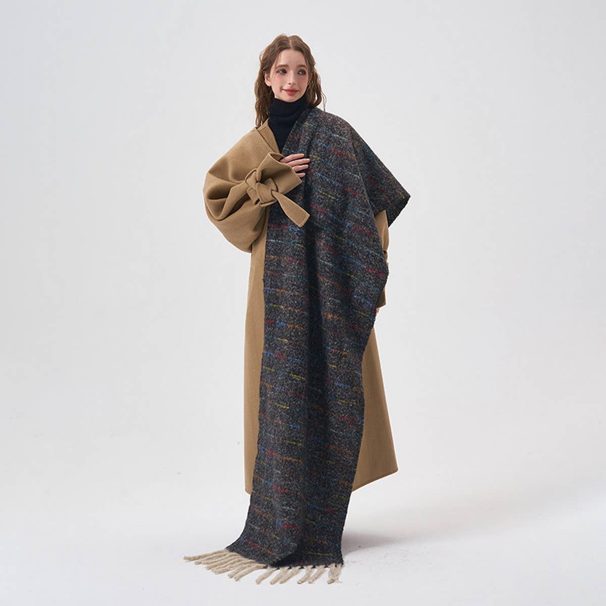 A woman models a long, double-layered wrap coat featuring a solid camel brown side and a textured multicolor tweed patterned side with cream fringe, worn over a black turtleneck against a plain white studio background.