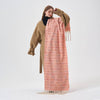 A model wearing a long, peach-pink woven scarf with thin multicolor horizontal stripes and cream fringe, styled over a tan coat and black turtleneck against a plain white studio background.