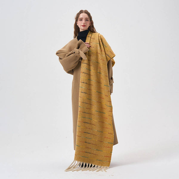 A woman modeling a long mustard yellow knit scarf with horizontal textured stripes and fringe, styled over a tan coat and black turtleneck against a plain white studio background.