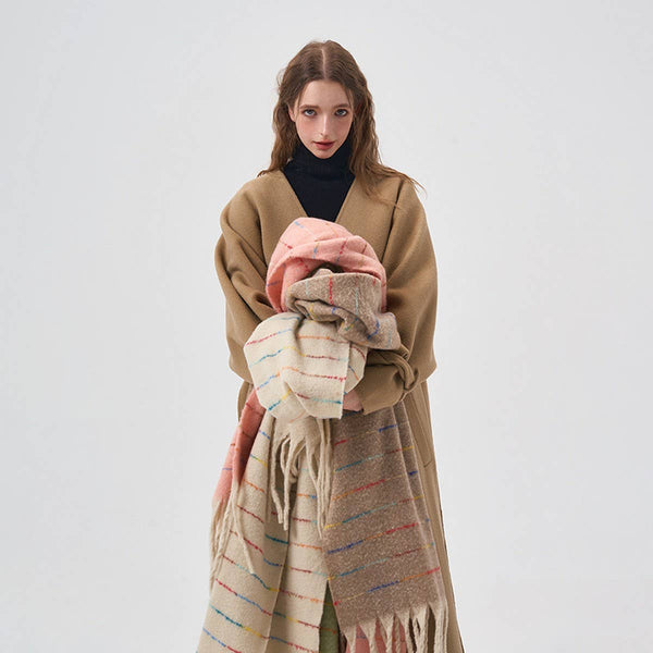A woman stands against a light gray studio background wearing a camel-colored wrap coat over a black turtleneck, while holding a soft beige scarf with thin multicolor stripes and a pink shawl.