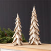 Two decorative tree sculptures in a metallic gold finish, displayed on a light wood surface with green pine branches against a dark gray background.