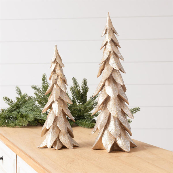 Two decorative metallic gold Christmas tree ornaments of different sizes displayed on a wooden table with green pine branches in the background.