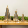 Three gold-toned metal Christmas tree ornaments with textured silver glass panels, displayed on a wooden shelf with green pine garlands against a light beige wall.