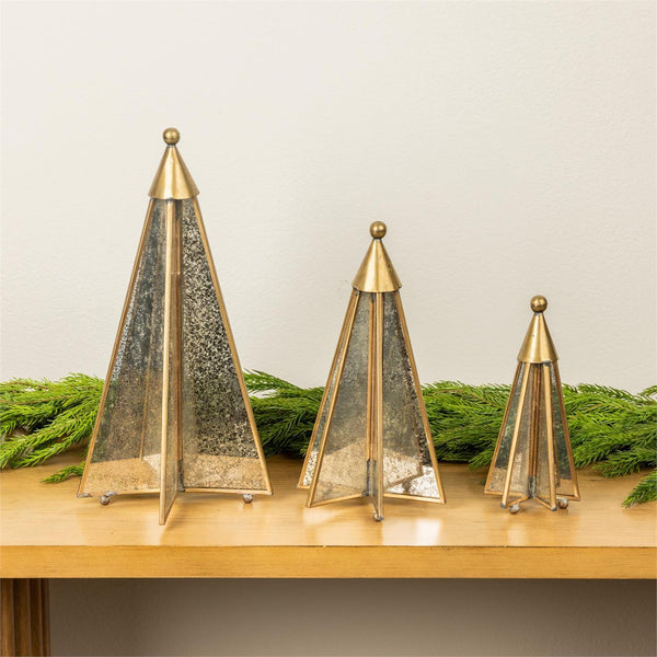 Three gold-toned metal Christmas tree ornaments with textured silver glass panels, displayed on a wooden shelf with green pine garlands against a light beige wall.