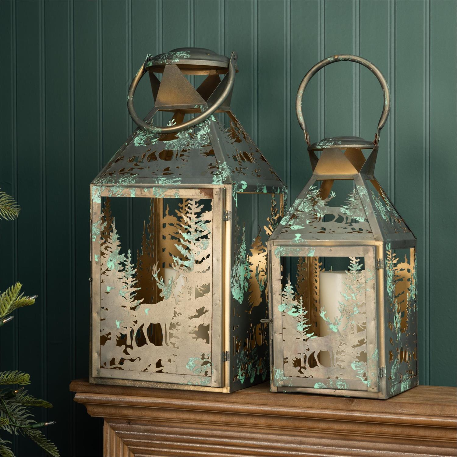 Two decorative metal lanterns with winter forest cutouts and hanging loops, displayed on a wooden surface against a dark green paneled wall.