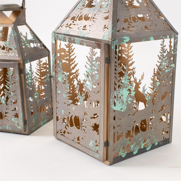 Decorative metal lanterns with a forest silhouette cutout design, featuring a weathered bronze finish with distressed turquoise patina accents, shown on a white background.