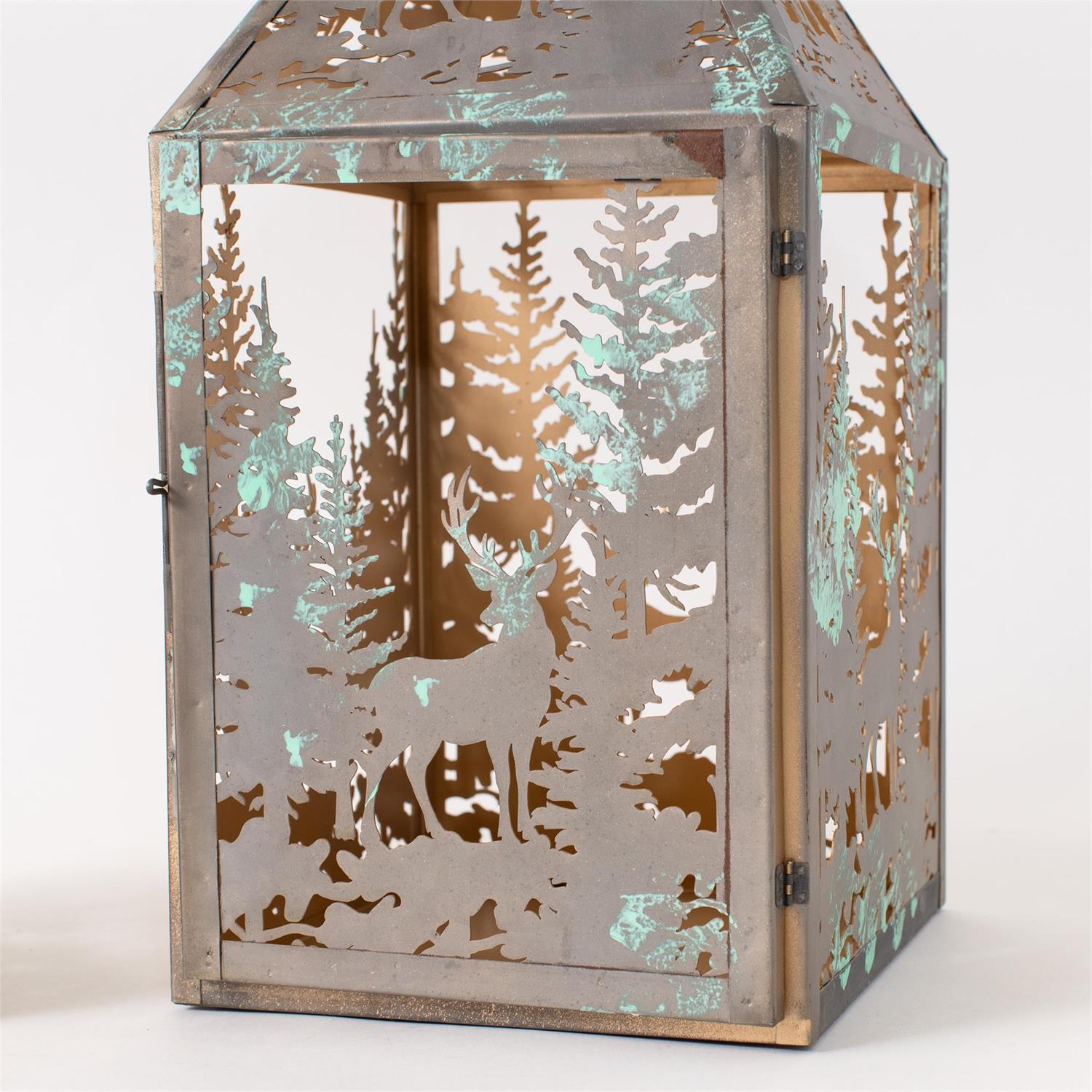 A decorative metal lantern with a forest silhouette cutout design, featuring a weathered silver-gray finish with turquoise patina accents and warm gold-toned interior lighting visible through the cutouts.
