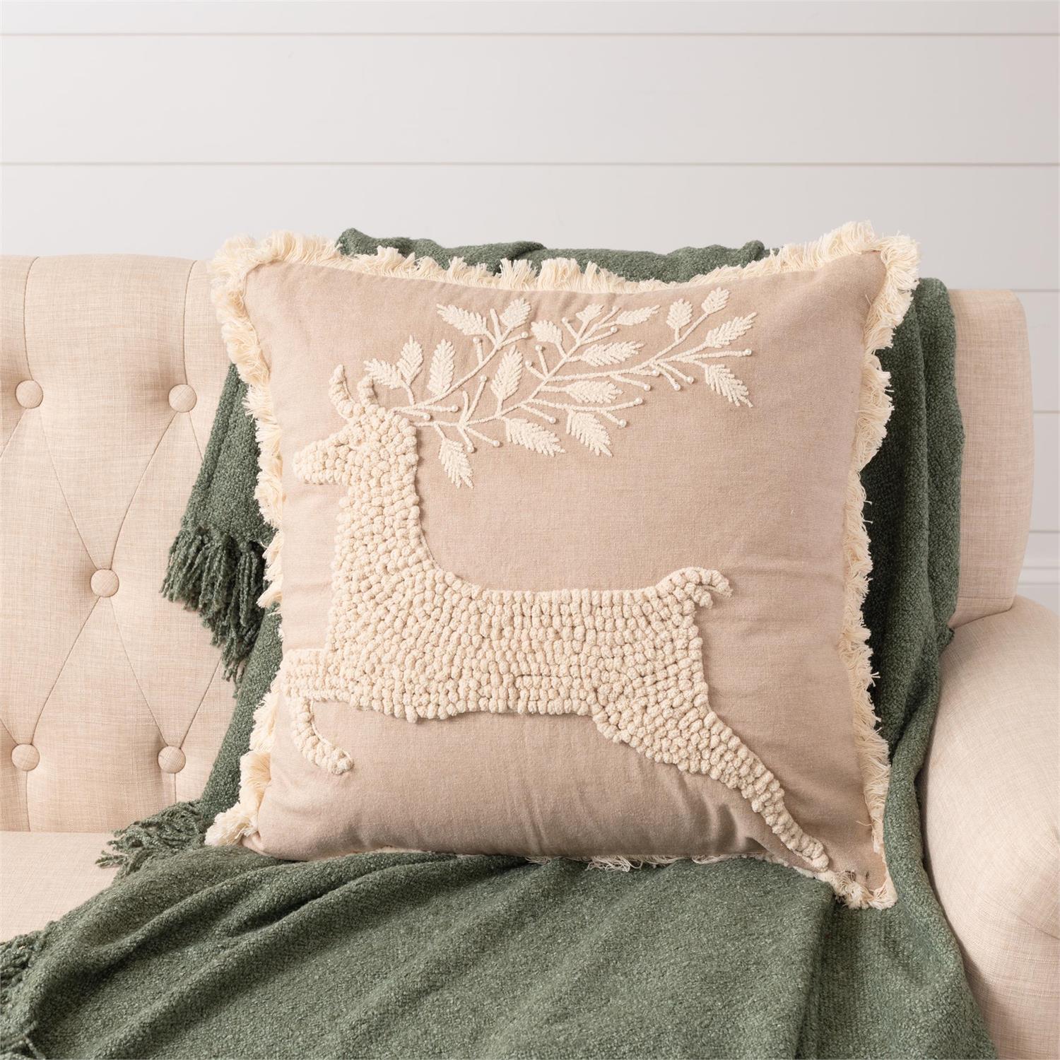 A square beige throw pillow with a raised cream beaded deer and leaf design, resting on a tufted beige armchair with a green throw blanket behind it.