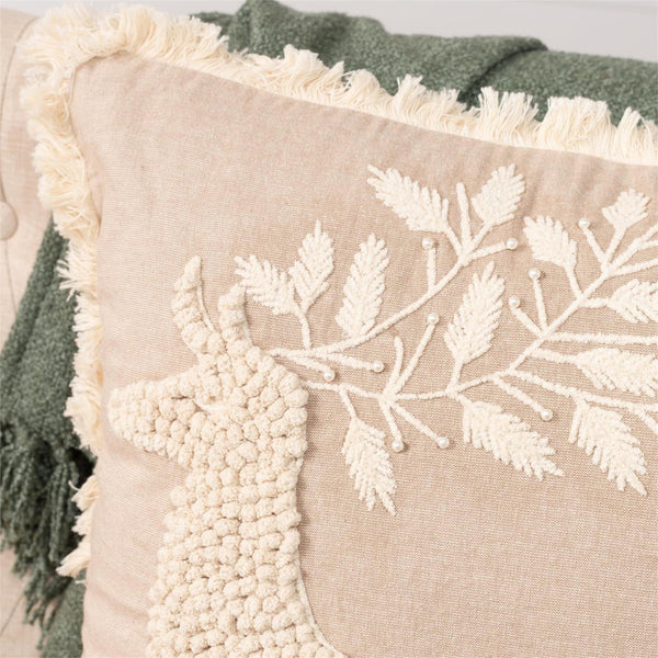 A close-up of a beige throw pillow with cream-colored embroidered leafy branches and a textured pom-pom deer motif, styled with a green fringed throw blanket.