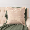 A square beige throw pillow with a raised cream beaded deer and leaf design, resting on a tufted beige armchair with a green throw blanket behind it.