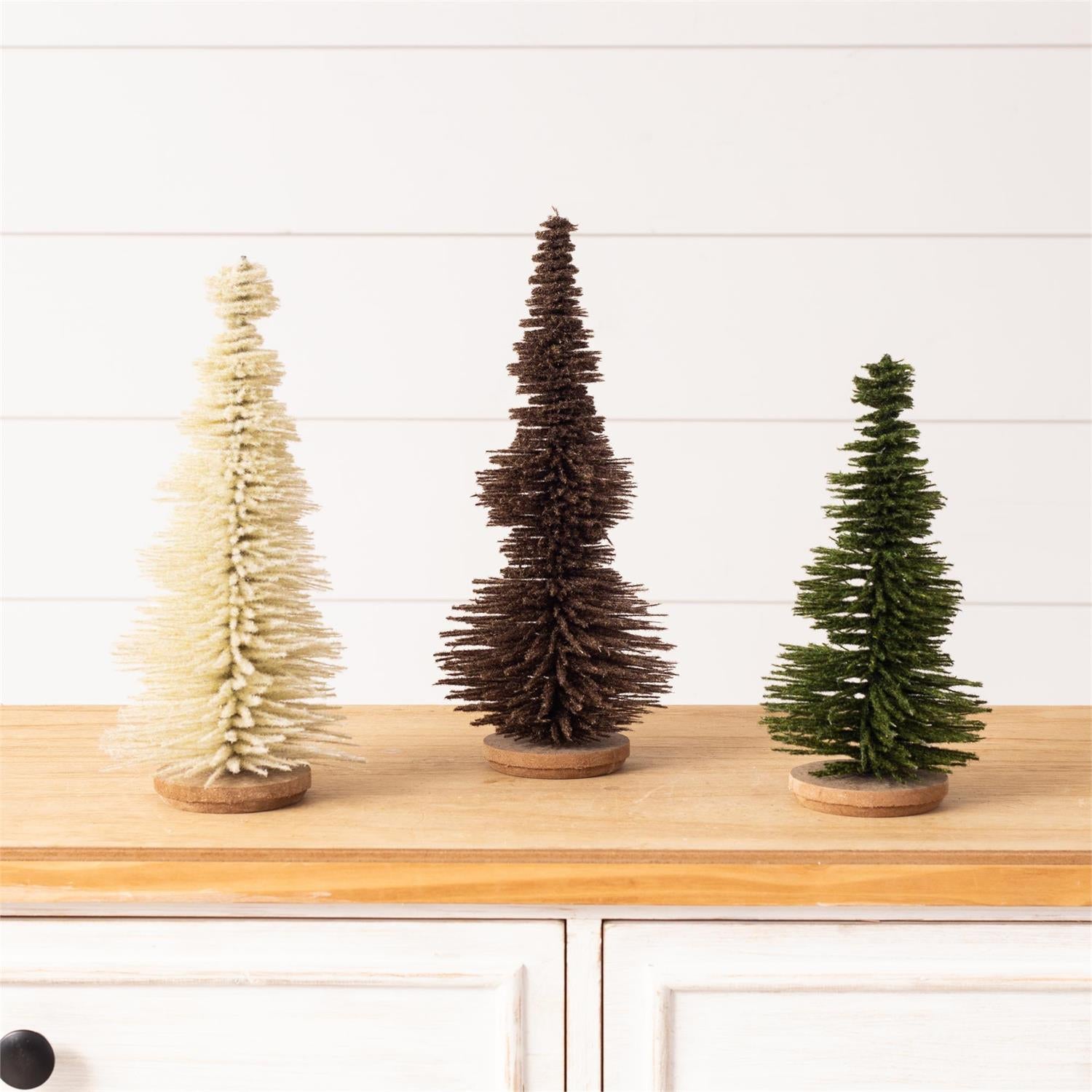 Three small decorative Christmas trees in cream, dark brown, and green, displayed on a light wood surface against a white paneled wall.