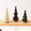 Three small decorative Christmas trees in cream, dark brown, and green, displayed on a light wood surface against a white paneled wall.