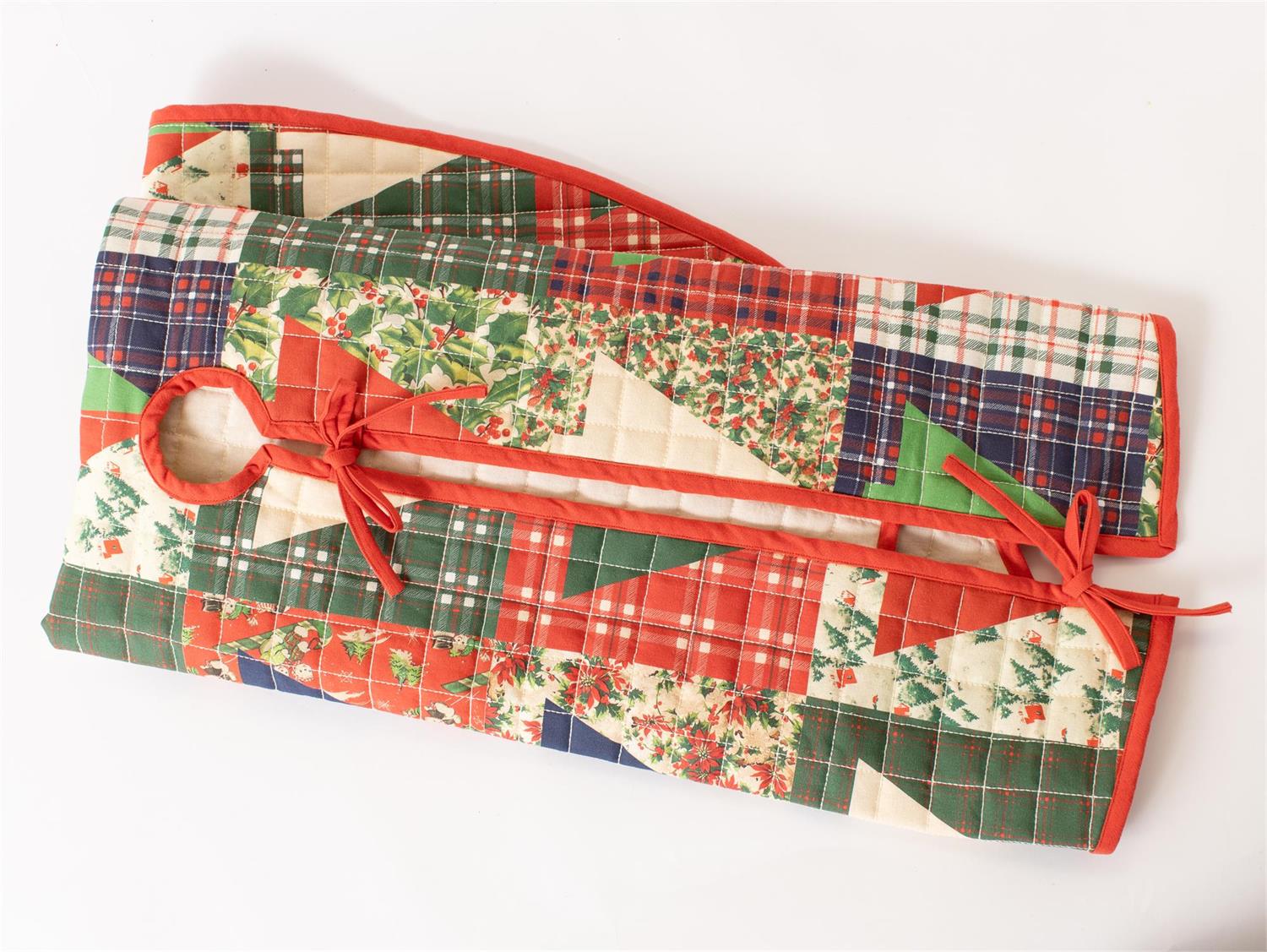 A quilted Christmas-themed quilted bag or organizer with a patchwork design of red, green, and beige fabrics, featuring a holly and pine pattern, plaid sections, and red ties, shown on a plain white background.