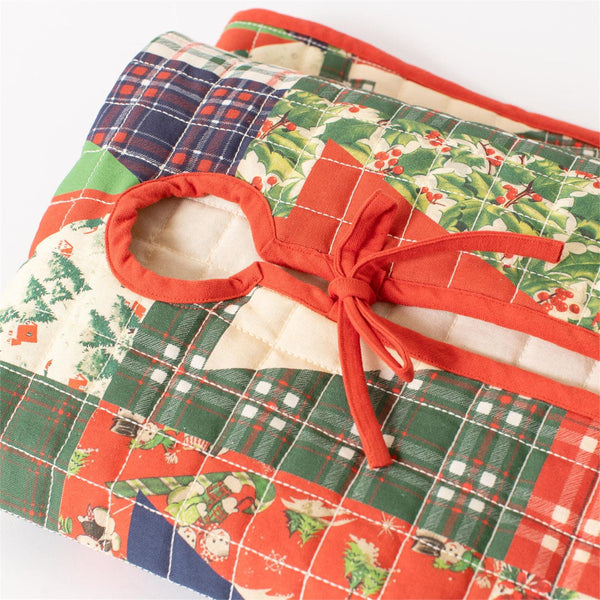 A close-up of a quilted fabric item, likely a quilted bag or pouch, featuring a festive Christmas patchwork design with red, green, and blue plaid, holly and pine patterns, and a red fabric bow detail on a white background.