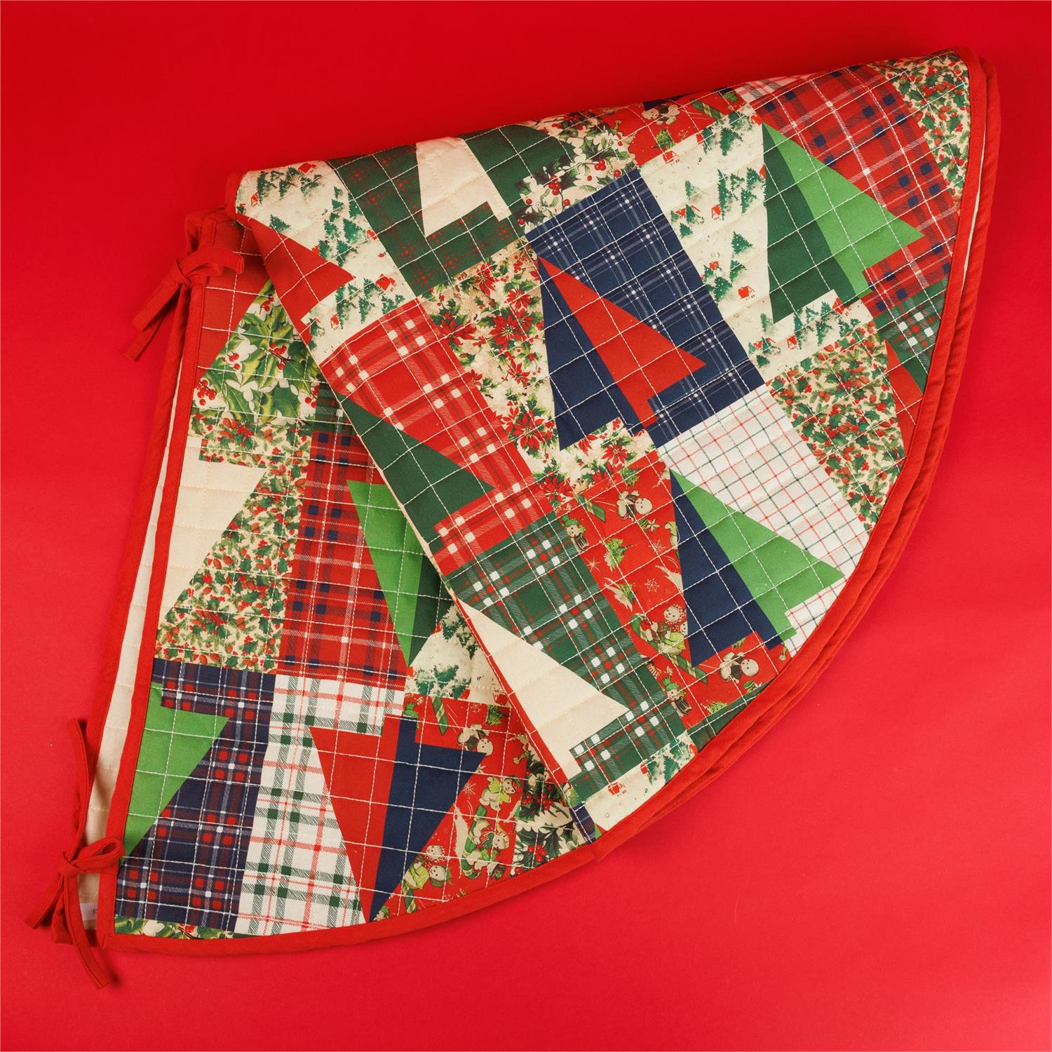 A Christmas-themed quarter-circle tree skirt with a patchwork design of red, green, navy, and cream fabrics, featuring plaid, floral, and geometric patterns, set against a solid red background.