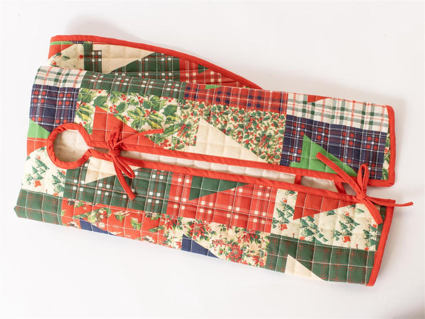 A quilted Christmas-themed quilted bag or organizer with a patchwork design of red, green, and beige fabrics, featuring a holly and pine pattern, plaid sections, and red ties, shown on a plain white background.