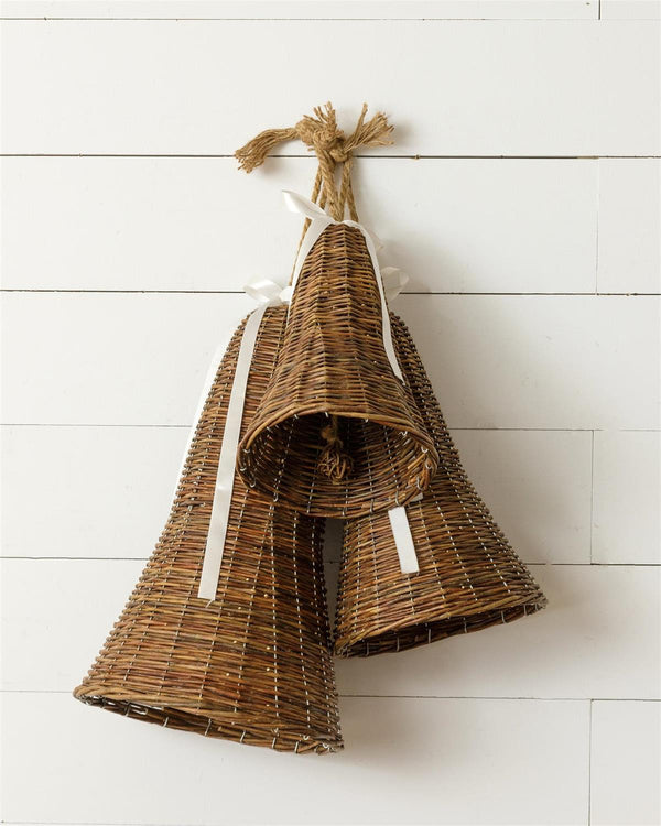Two woven wicker hanging bells in natural brown tones, tied together with white ribbons and suspended from a jute rope against a white wood plank wall.