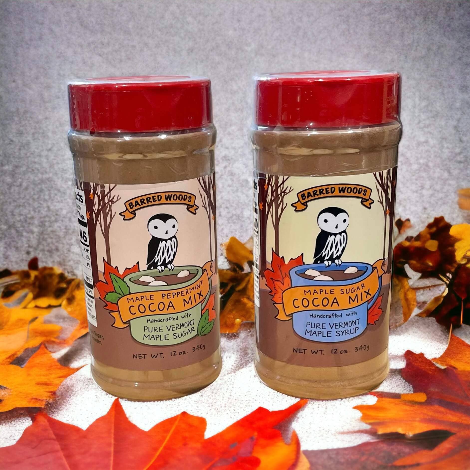 Two jars of Barred Woods Maple Peppermint Cocoa Mix and Maple Sugar Cocoa Mix, displayed side-by-side on a white surface surrounded by autumn leaves against a gray textured background.