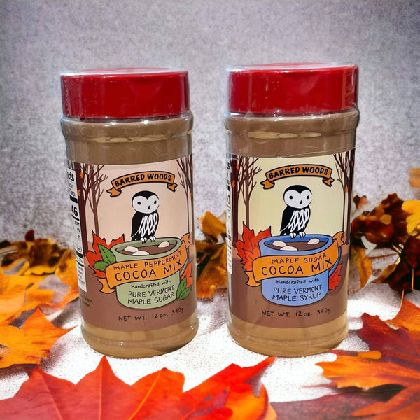 Two jars of Barred Woods Maple Peppermint Cocoa Mix and Maple Sugar Cocoa Mix, displayed side-by-side on a white surface surrounded by autumn leaves against a gray textured background.