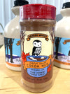 A jar of Barred Woods Maple Sugar Cocoa Mix in the foreground, with maple syrup bottles blurred in the background on a light wood surface.