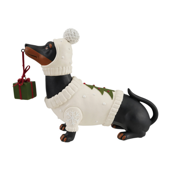 A dachshund Christmas ornament figurine wearing a white knit sweater and matching hat with a pom-pom, holding a small green gift box with a red ribbon. The figurine has a black and tan body with a white sweater featuring green pine tree accents.