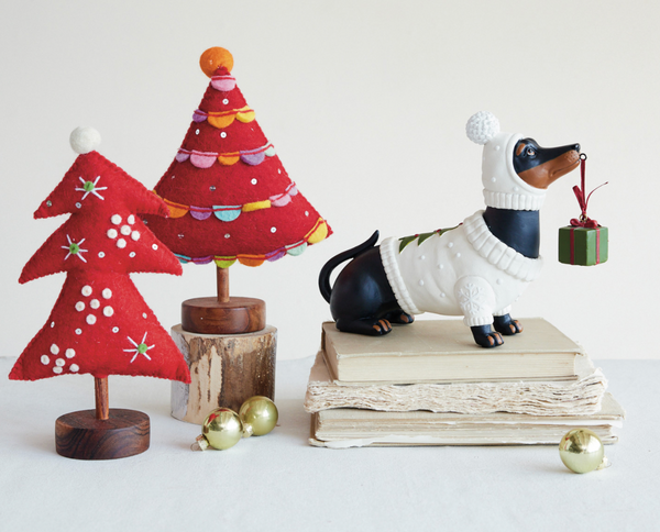 A styled holiday tabletop scene with two red felt Christmas trees on wooden bases, a black-and-white dachshund figurine wearing a white knit sweater and holding a small green gift box, stacked vintage books, and a few gold ornaments against a plain light background.