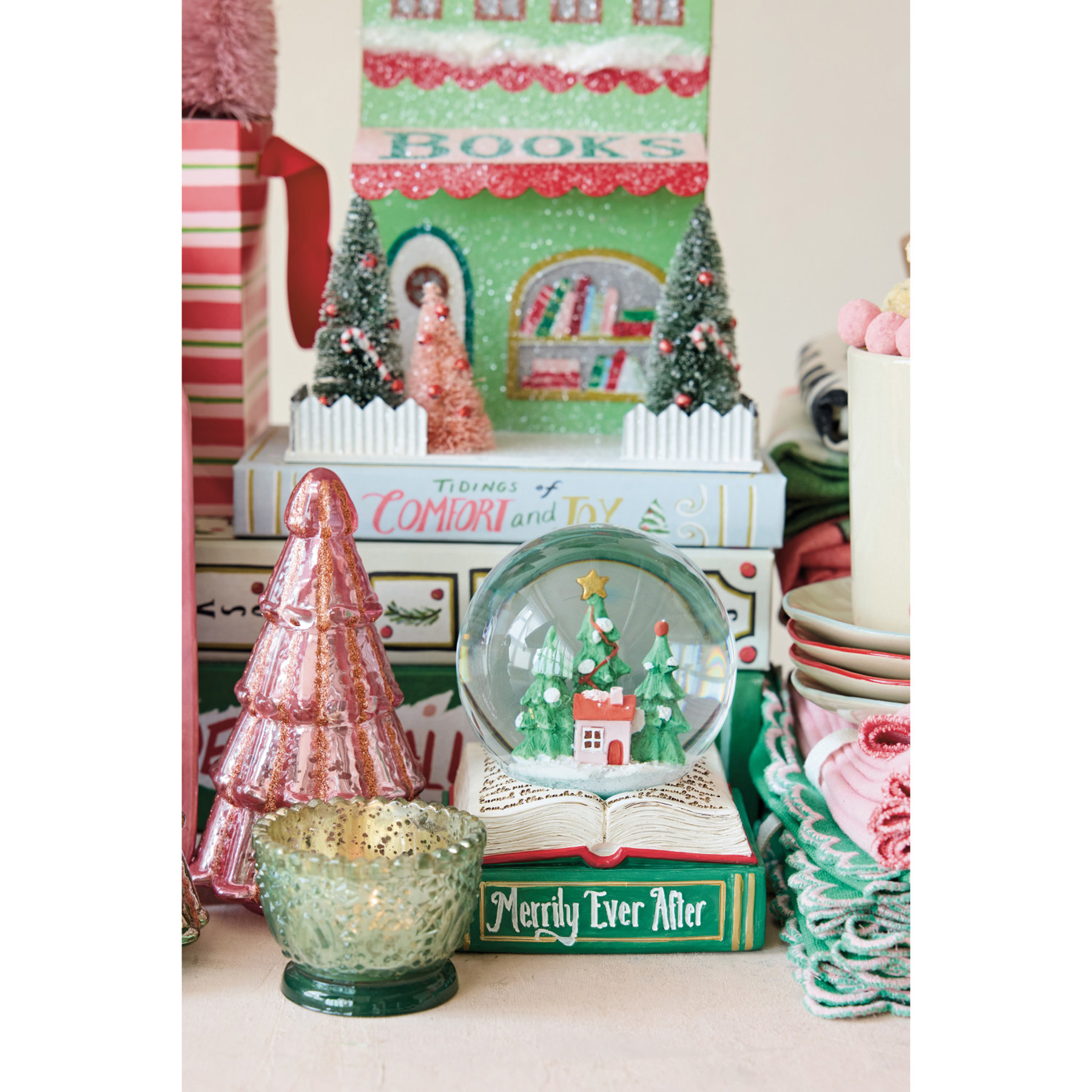 A collection of vintage Christmas-themed decorative items, including a green and red 'Books' book-shaped ornament, a pink glittery Christmas tree, a green glass candle holder, and a snow globe with a miniature Christmas village scene on an open book.