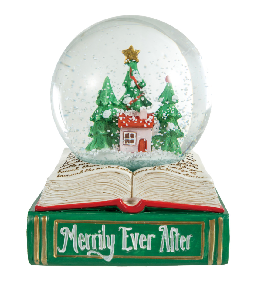 A Christmas-themed snow globe figurine resting on an open book base. The clear globe contains a miniature winter scene with green Christmas trees, a small house, snow, and a gold star.