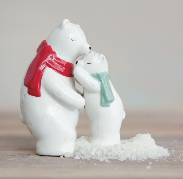 A small white ceramic polar bear figurine showing a mother bear hugging her cub, with the mother bear wearing a red scarf and the cub wearing a mint green scarf. They are standing on white decorative salt crystals on a light wooden surface against a soft beige background.