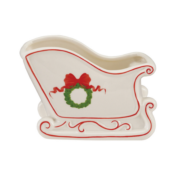 A white ceramic Santa's sleigh-shaped dish or planter with red decorative lines and a green wreath with a red bow on the front, shown on a plain white background.
