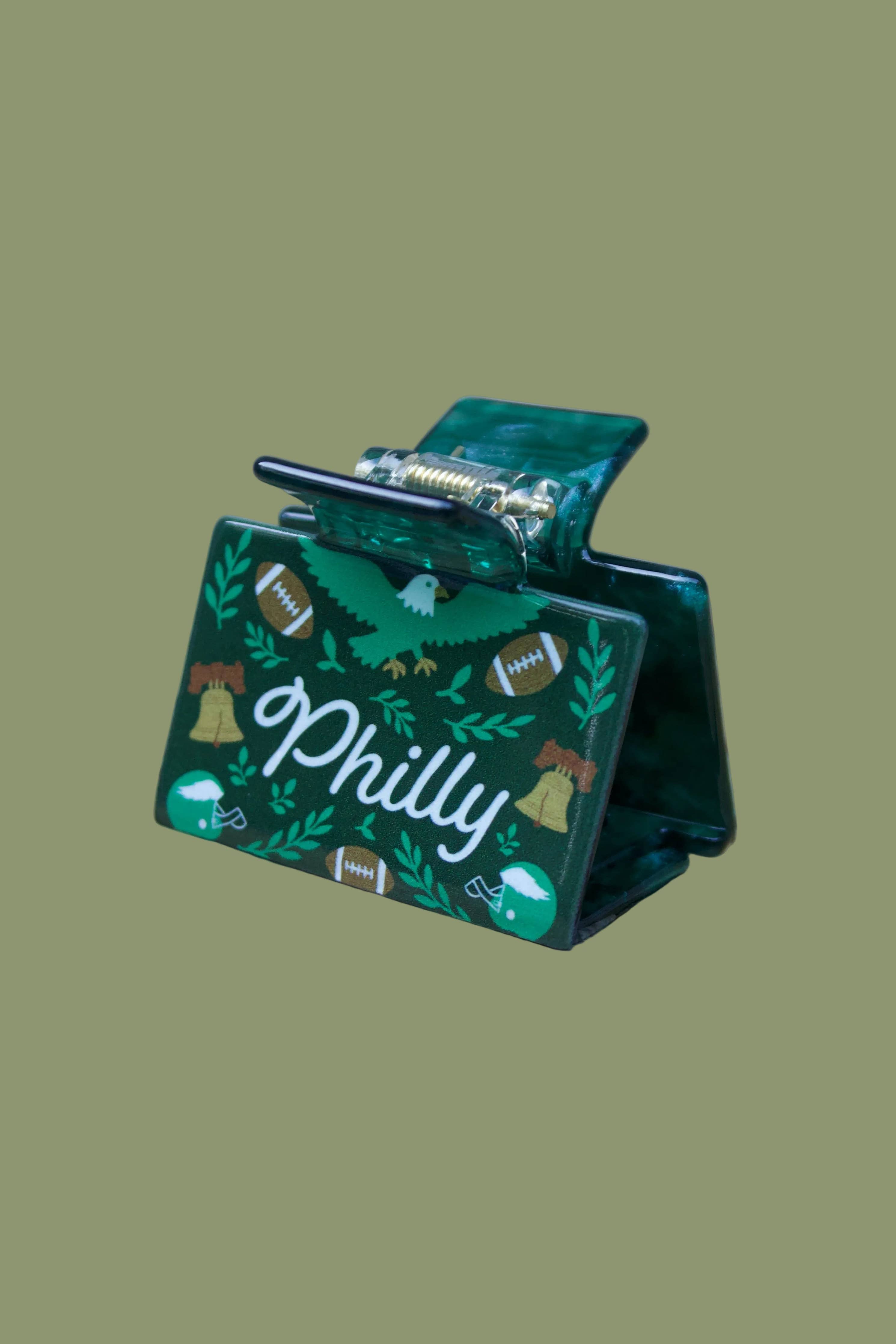 A green hair claw clip with a Philadelphia theme, featuring a hand-drawn eagle, footballs, bells, and the text 'Philly' in white script, resting on a wooden slatted surface.
