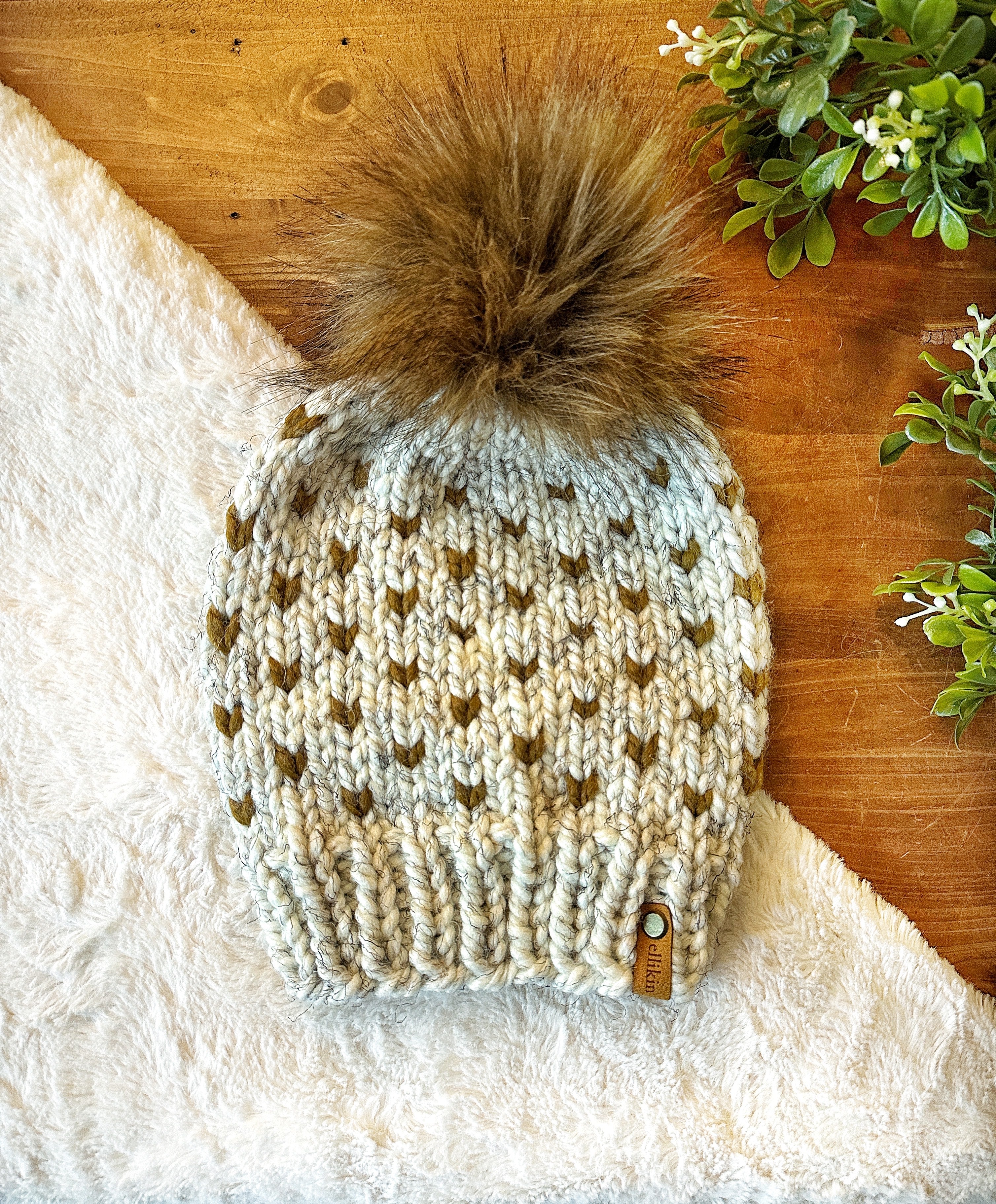 A chunky knit beanie hat with a large brown faux-fur pom-pom, laid flat on a white faux-fur surface over a wooden tabletop with small green leaves in the corner. The beanie is cream and olive green with a small tan brand tag near the brim.
