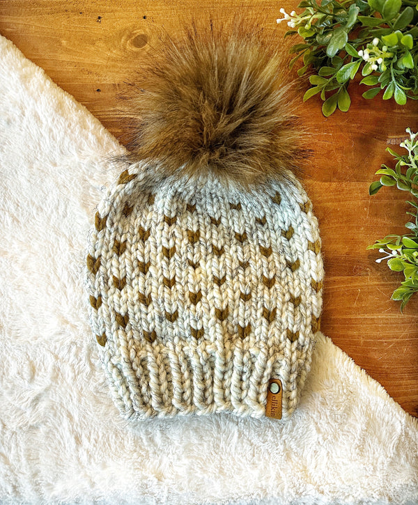 A chunky knit beanie hat with a large brown faux-fur pom-pom, laid flat on a white faux-fur surface over a wooden tabletop with small green leaves in the corner. The beanie is cream and olive green with a small tan brand tag near the brim.