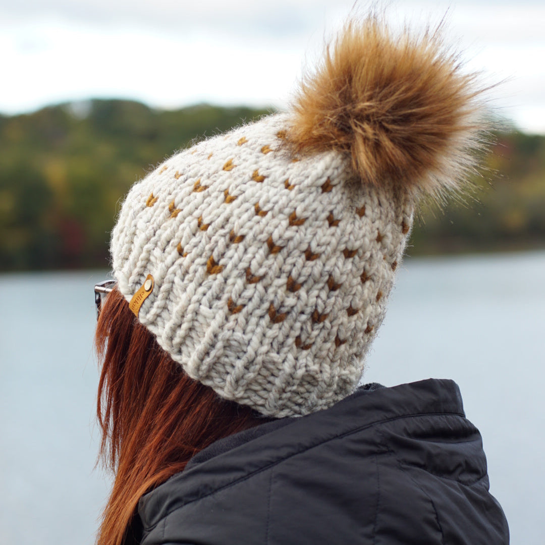 A person viewed from the back wearing a cream knit beanie with a large brown faux-fur pom-pom and small brown star-like knit details, outdoors near water with trees in the background.