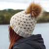 A person viewed from the back wearing a cream knit beanie with a large brown faux-fur pom-pom and small brown star-like knit details, outdoors near water with trees in the background.