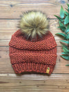 A chunky knit beanie in a warm rust orange color, featuring a large tan and brown faux fur pom-pom on top. The beanie is laid flat on a rustic wooden surface next to a green leafy sprig.
