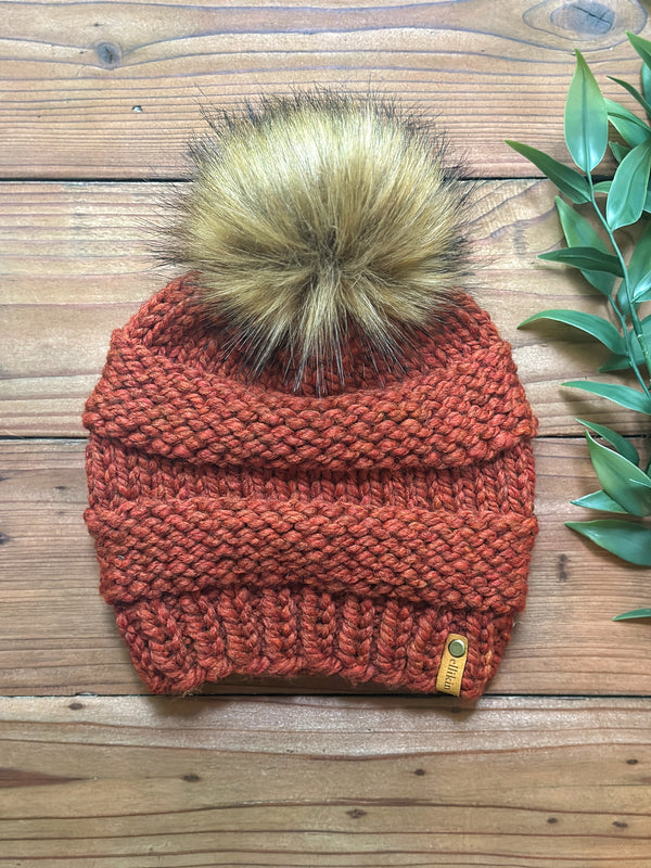 A chunky knit beanie in a warm rust orange color, featuring a large tan and brown faux fur pom-pom on top. The beanie is laid flat on a rustic wooden surface next to a green leafy sprig.