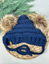 Blue crocheted beanie with fur pom-poms on a woven surface with greenery.