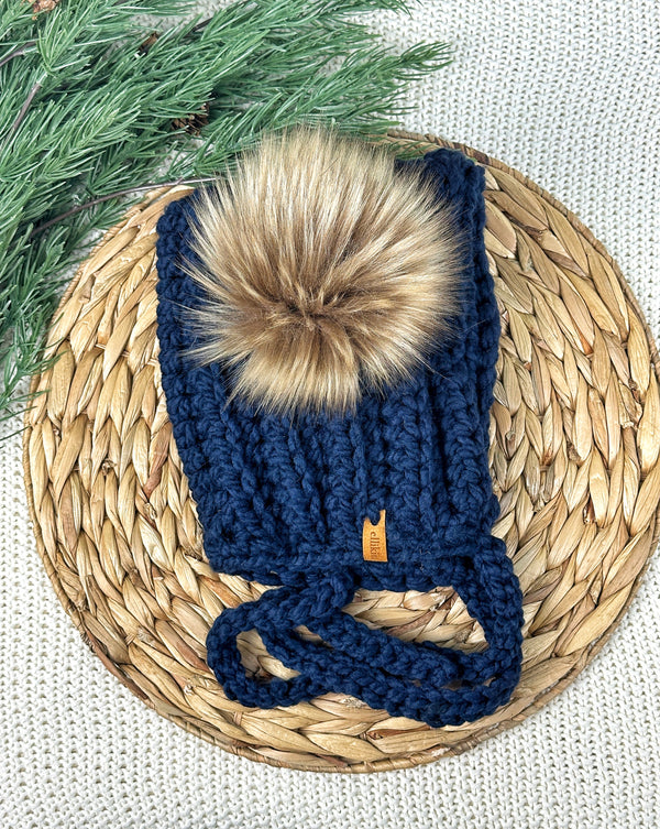 A navy blue crocheted beanie hat with a large tan faux-fur pom-pom and a small tan leather logo tab, displayed on a round woven straw mat with green pine branches in the corner.