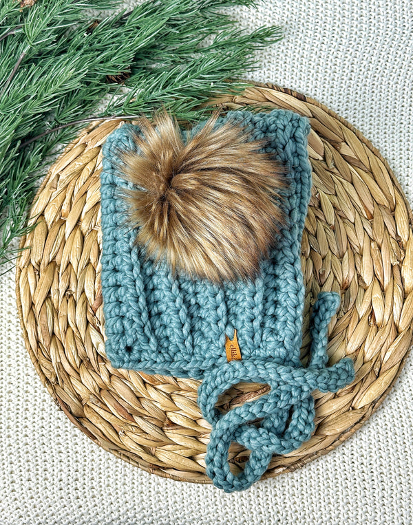 Knitted blue baby hat with a pom-pom on a woven mat