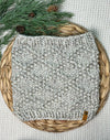 A light gray heathered knit cowl or neck warmer with a small brown leather brand tab, displayed on a round woven straw mat with pine branches and pinecones.