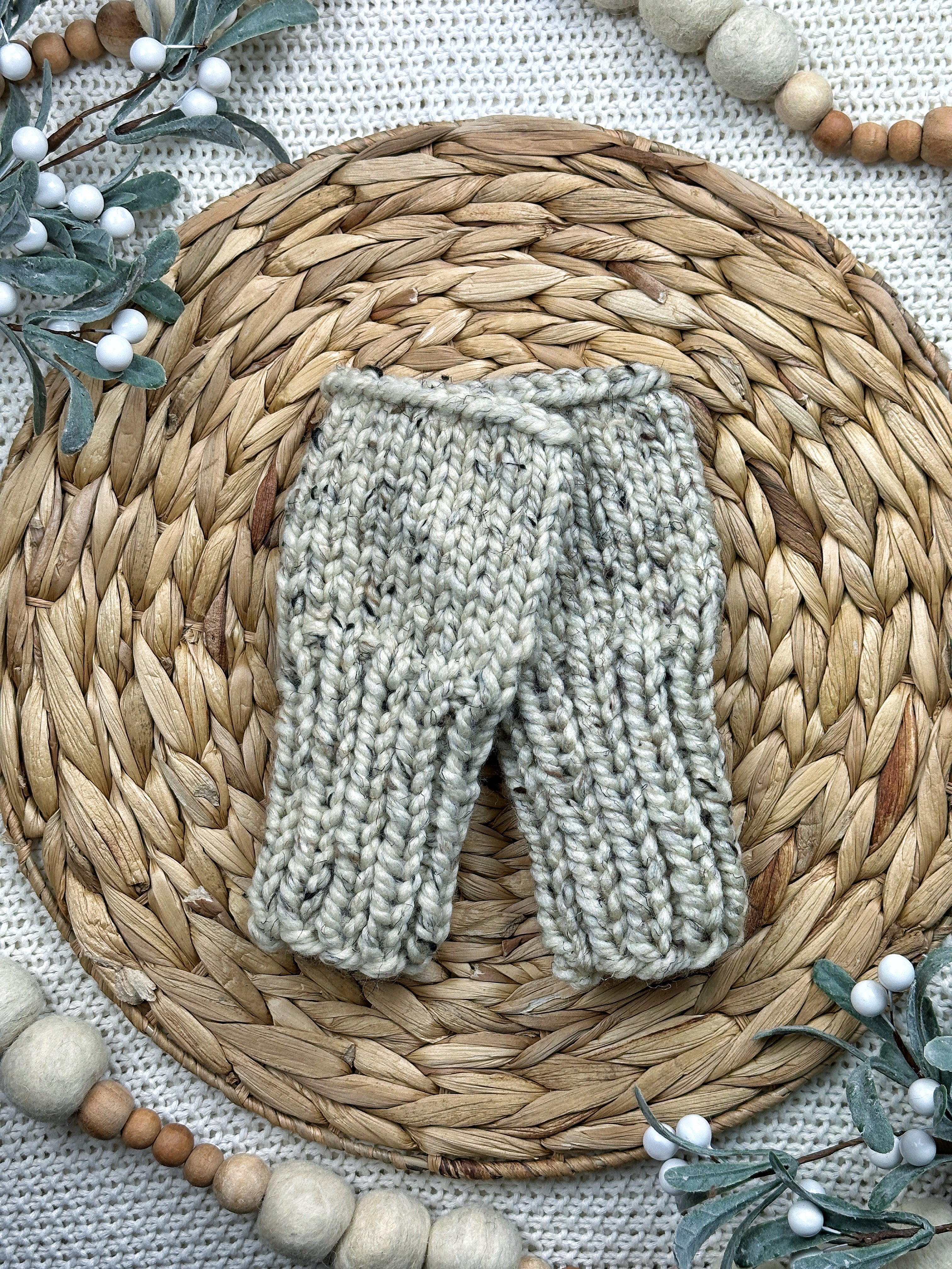 A pair of hand-knit baby booties in a light oatmeal gray color, displayed on a round woven straw placemat with decorative wooden beads and eucalyptus branches.