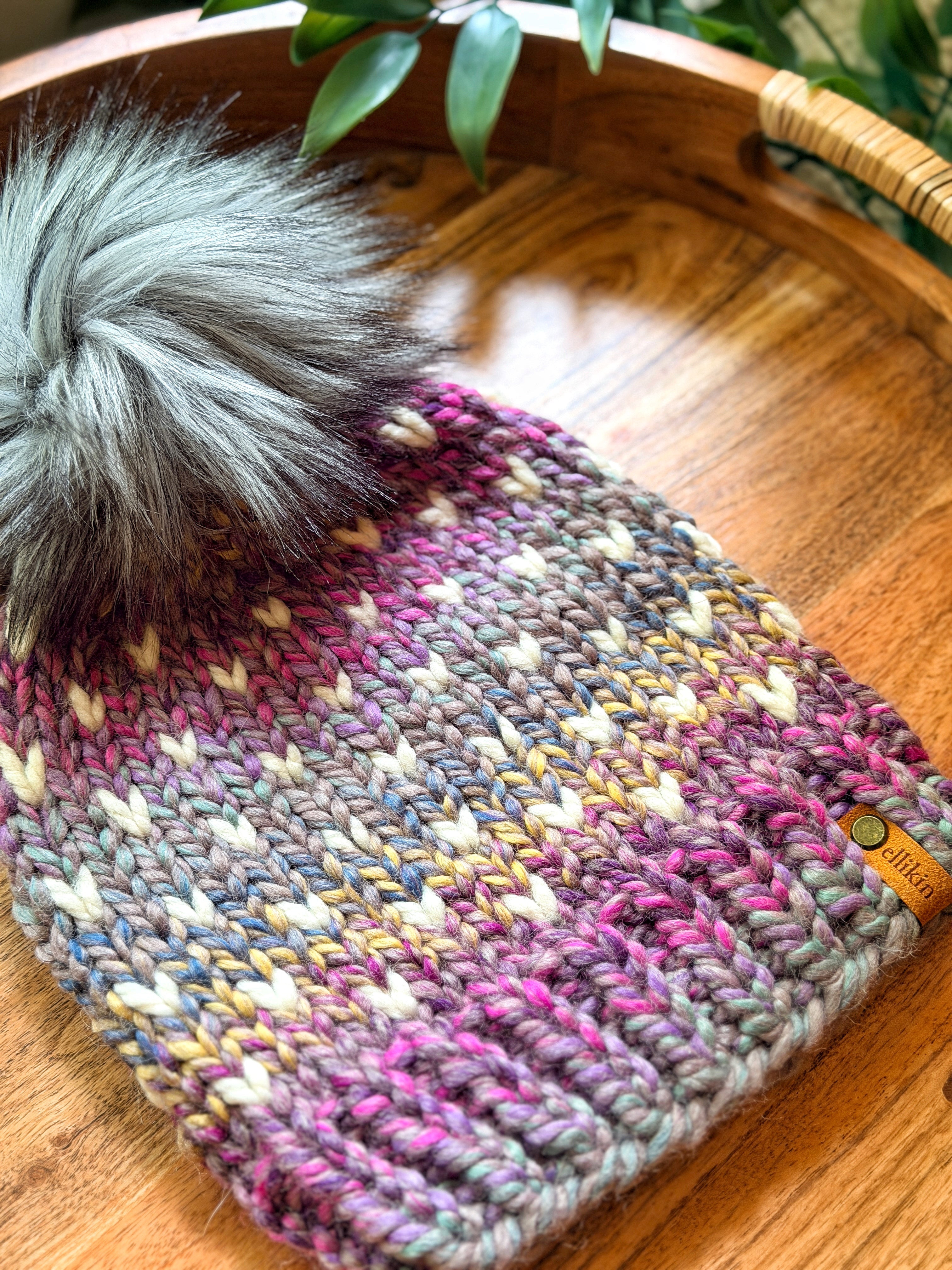 A chunky knit beanie hat with a large gray faux-fur pom-pom, featuring a multicolor variegated pattern of pink, purple, blue, yellow, and white. A small tan leather brand tag is visible on the side, resting on a wooden tray.