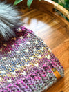 A close-up of a chunky knit beanie hat made from variegated yarn in pink, purple, gray, blue, and cream, featuring a fluffy gray faux fur pom-pom and a small tan leather tag that reads 'silkyn'. The hat is styled on a round wooden tray with green leaves in the background.
