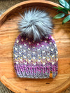 A hand-knit winter beanie with a large fluffy gray faux-fur pom-pom, featuring a multicolor chevron pattern in pink, purple, blue, and cream. The beanie is displayed on a round wooden tray with a small leather brand tag on the side.