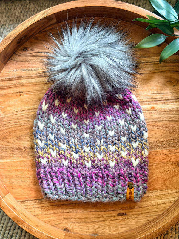 A hand-knit winter beanie with a large fluffy gray faux-fur pom-pom, featuring a multicolor chevron pattern in pink, purple, blue, and cream. The beanie is displayed on a round wooden tray with a small leather brand tag on the side.
