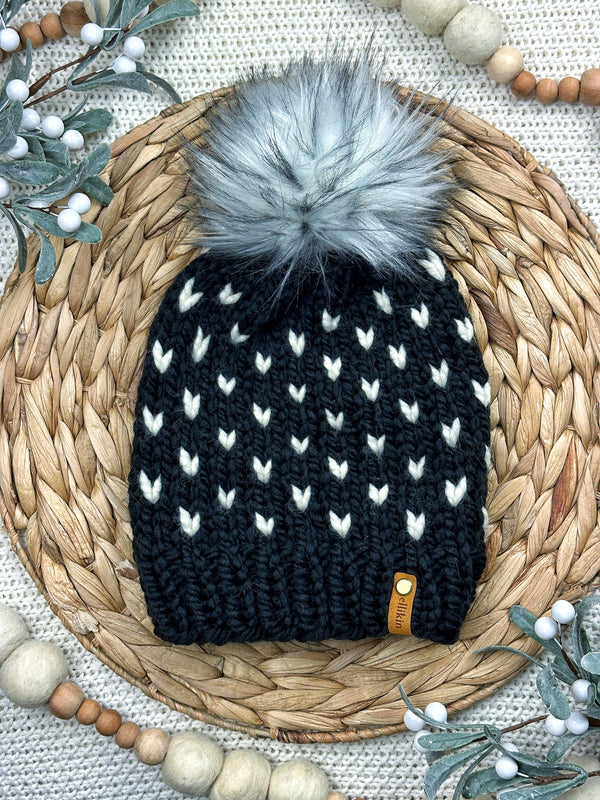 A black knit beanie with a white heart pattern and a large gray faux fur pom-pom, displayed on a woven straw mat with decorative wooden beads and greenery.